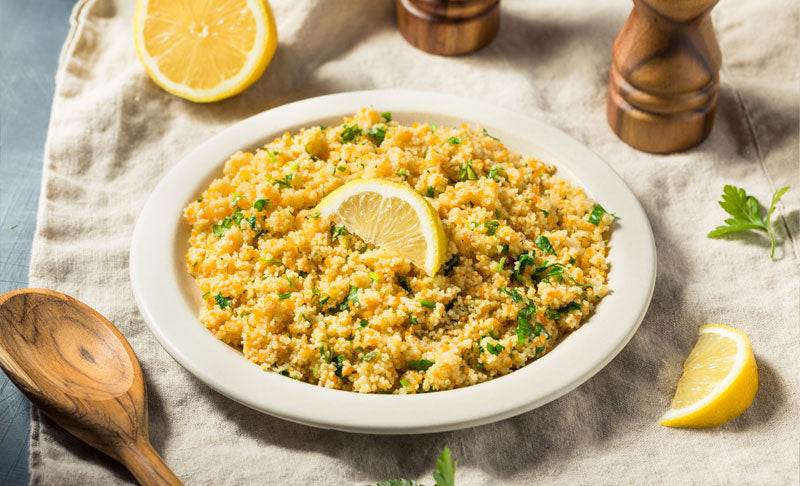Ein Teller CousCous mit Kräutern, Olivenöl und Zitrone auf einem dekorativen Leintuch, umgeben von Pfeffer und Salzmühle, ein paar Zitronenscheiben und Kräutern.
