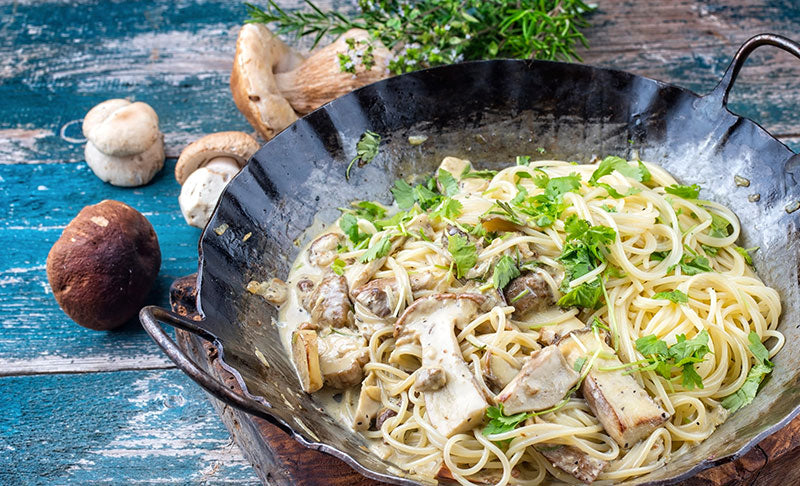 Tagliatelle mit Funghi Porcini in einem gußeisernen Pfännchen mit Griffen auf einem türkis bemalten Holztisch. Dekoriert mit einigen Pilzen und Kräutern